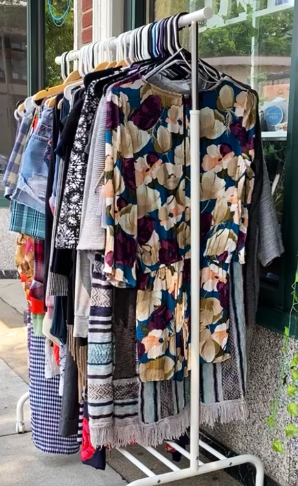 A clothing rack sitting outside the Uvida Shop Brookline location as part of our summer 2023 clothing swap. The rack is full of hangers with brightly colored and patterned clothes.